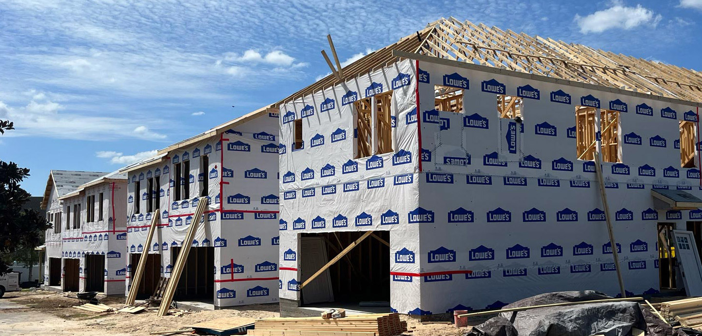 Under-construction multifamily building covered with Lowes-branded insulation wrap, wooden roof framing visible, clear sky above