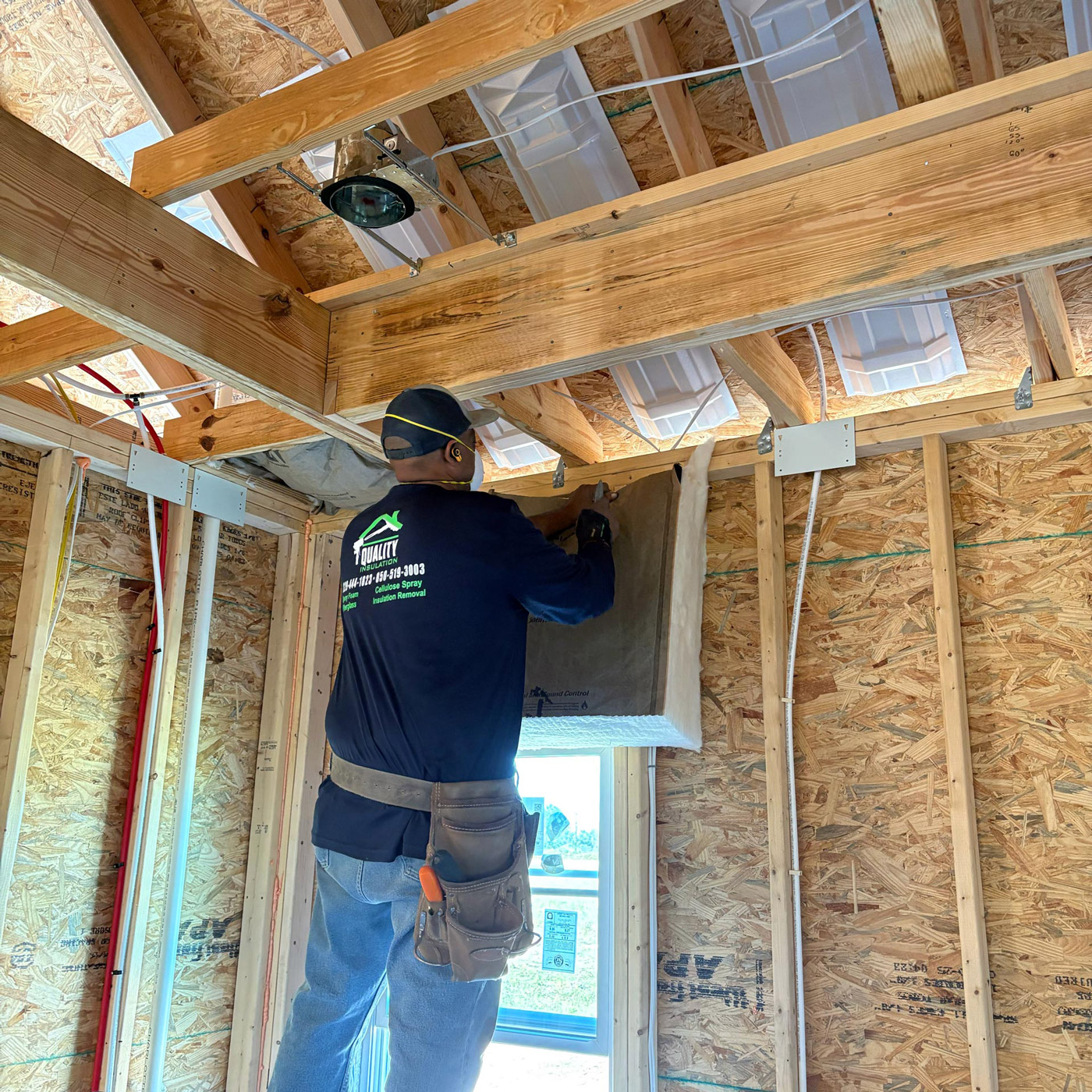 new-home-insulation Person installing insulation in unfinished house interior, wearing a tool belt and protective gear
