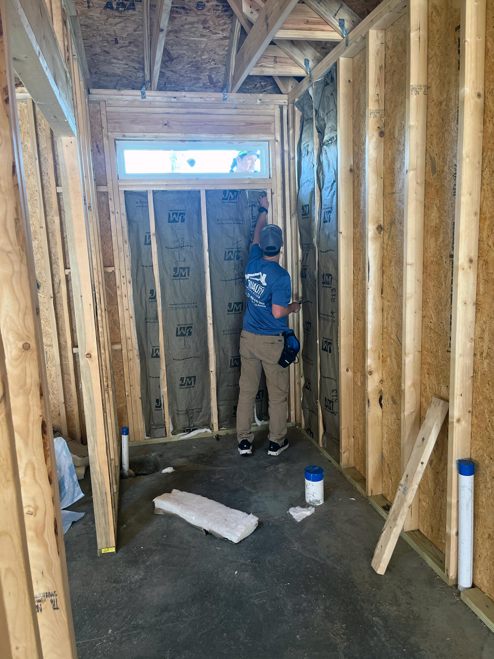 Technician installing fiberglass insulation in unfinished room, wearing blue shirt and cap