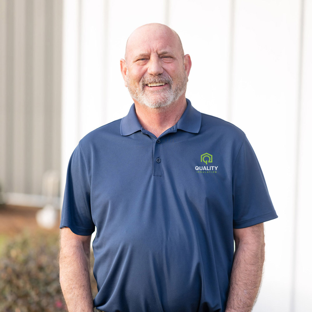 Man smiling outdoors in front of a building, wearing a navy polo with "Quality Insulation" logo