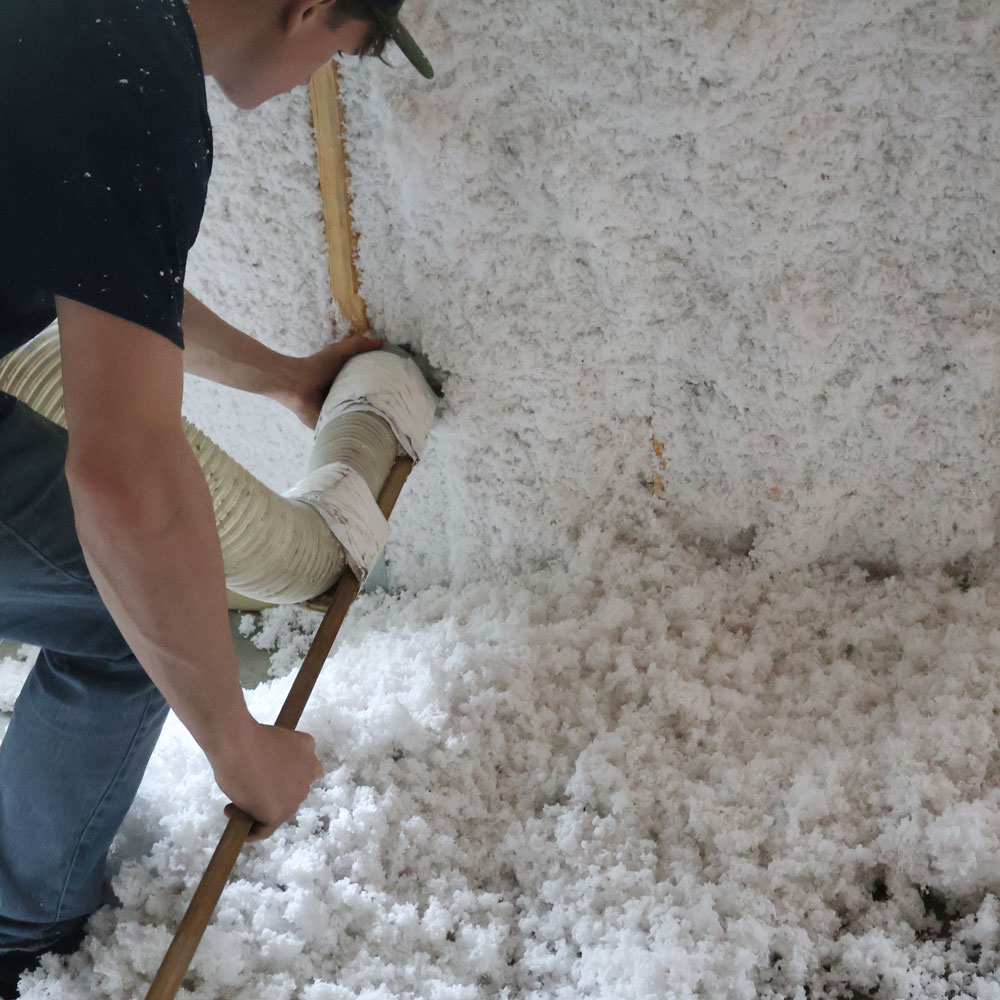 A person using a hose to install white fluffy insulation on a wall and floor