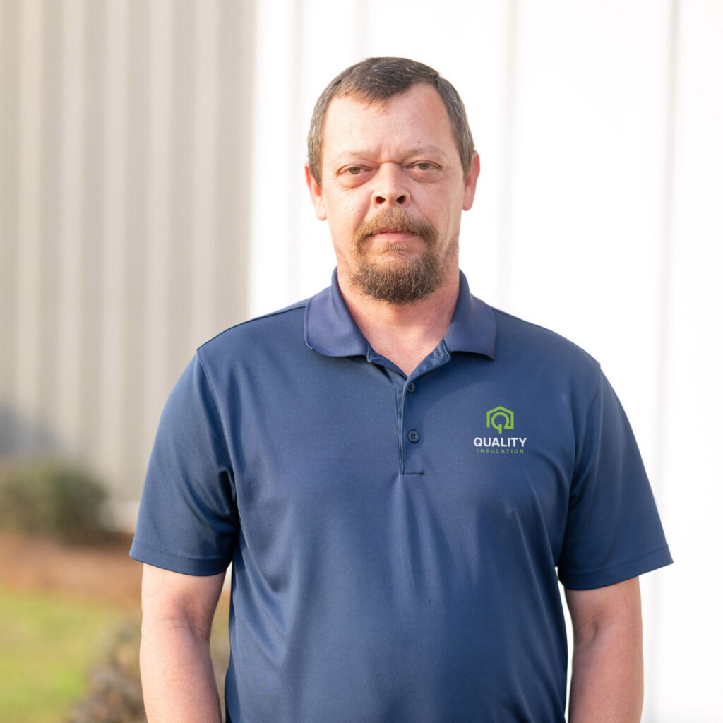 Man with beard wearing a blue "Quality Insulation" polo shirt stands outdoors facing the camera