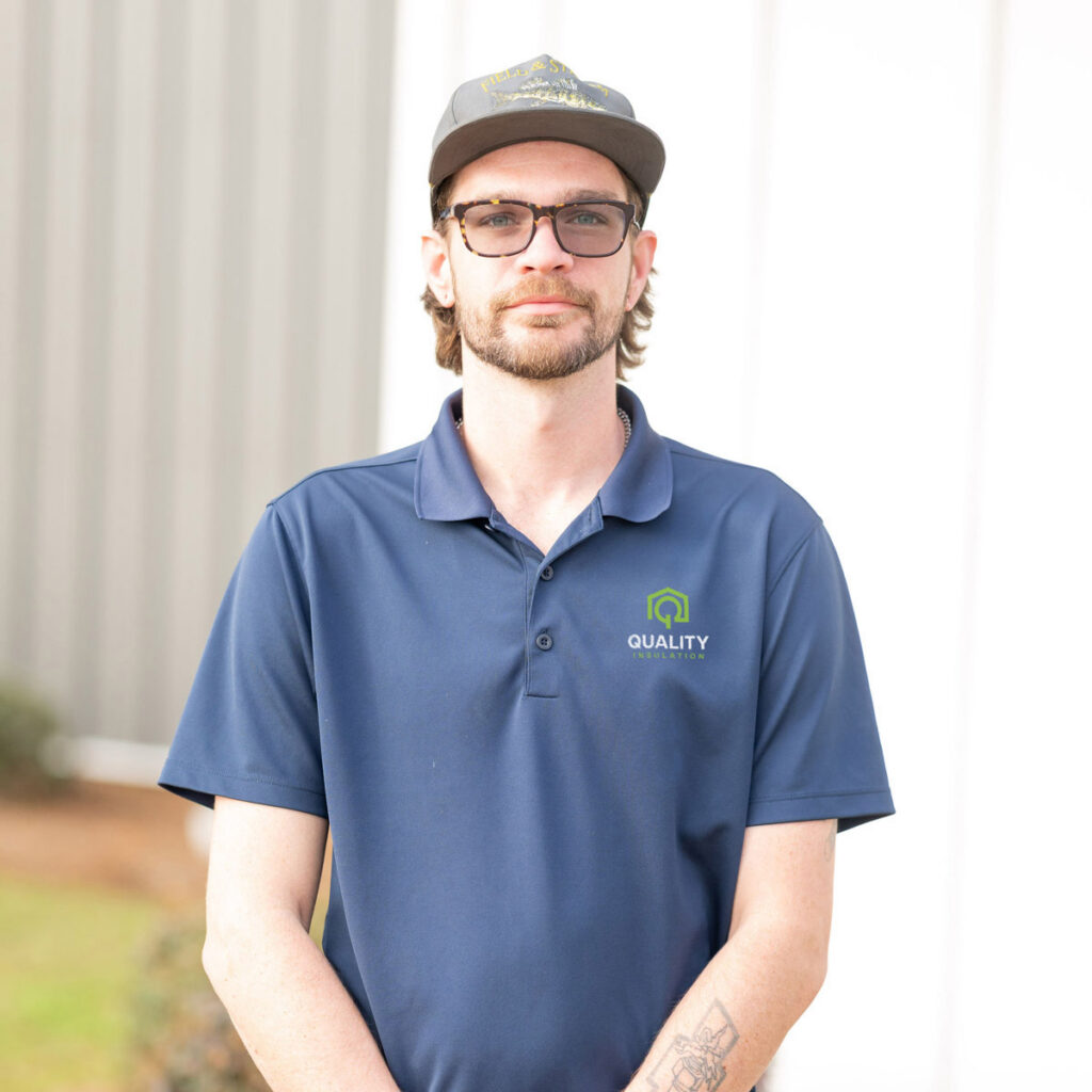 Man in blue "Quality Insulation" polo stands outside, wearing glasses and a cap