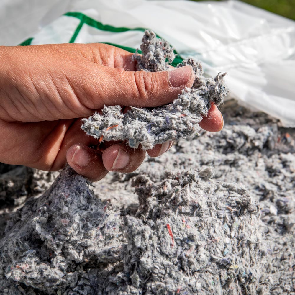 persons-hand-feeling-cellulose-insulation Hand holding clump of gray cellulose insulation over a larger pile, with a white and green bag in the background