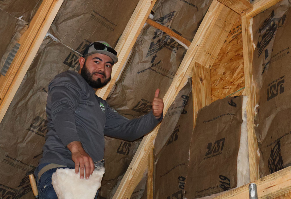 professional-insulation-installer Man installing insulation in attic gives thumbs up, surrounded by wooden beams and insulation material